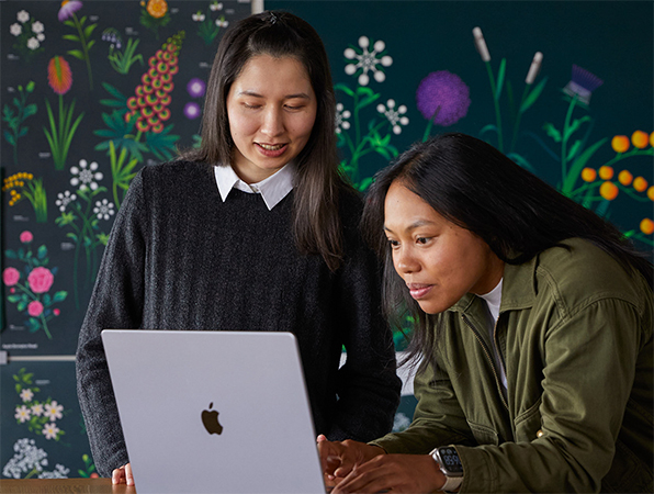 To ansatte p? Apples markedsf?ringsteam samarbeider p? en MacBook foran plakater designet av teamet.