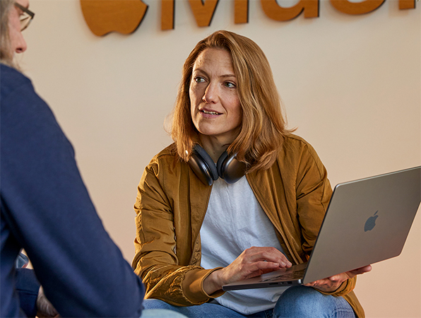 To ansatte p? Apples team for salg og forretningsutvikling snakker sammen. Den ene holder en MacBook og sitter foran en vegg med en Apple?Music-logo.