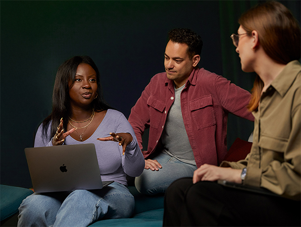Tre ansatte p? Apples team for programvare og tjenester sitter i en sofa og snakker sammen, den ene med en MacBook.