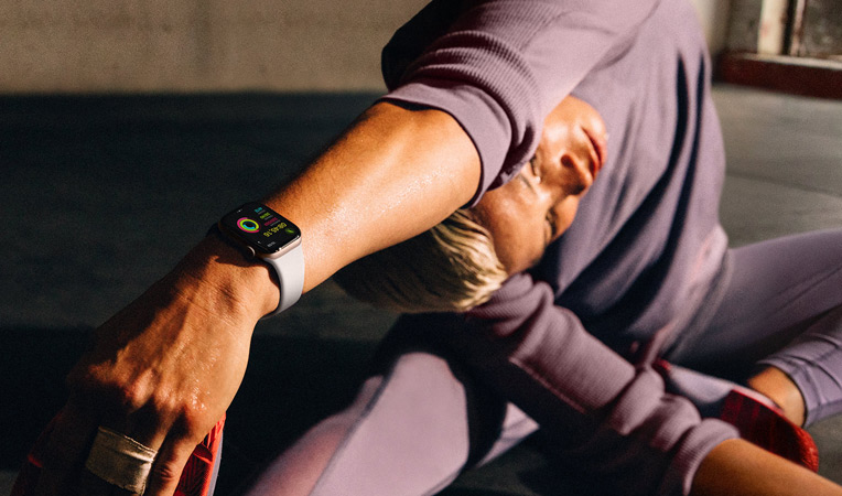 Eine Frau auf dem Boden dehnt ihren Arm zum Bein hin, w?hrend sie eine Apple Watch Series 10 tr?gt. Display-Inhalt: Training, Cooldown, Aktivit?t und Aktivit?tsringe