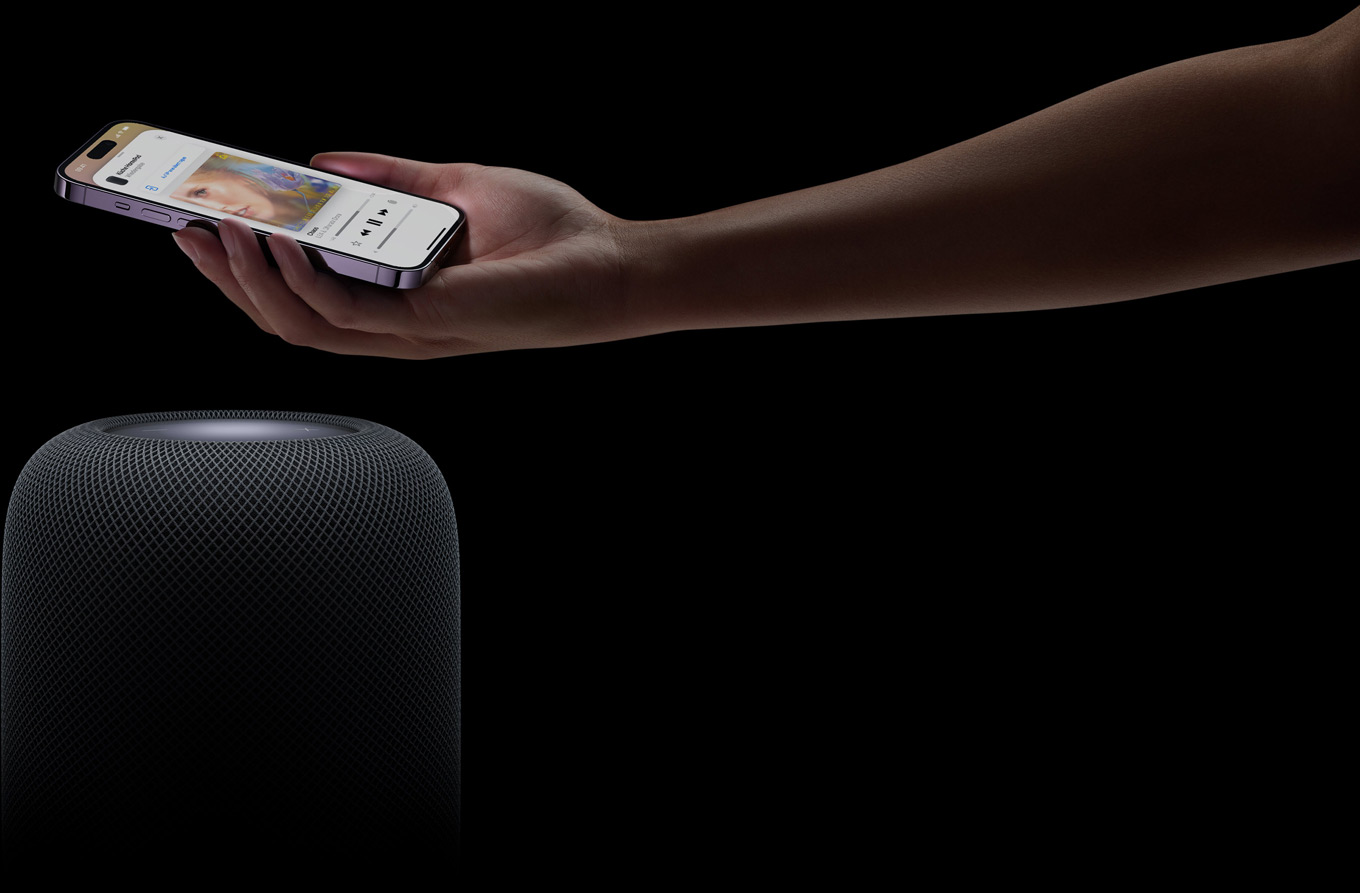 Eine Hand, die von rechts kommt, h?lt ein iPhone über einen HomePod Lautsprecher