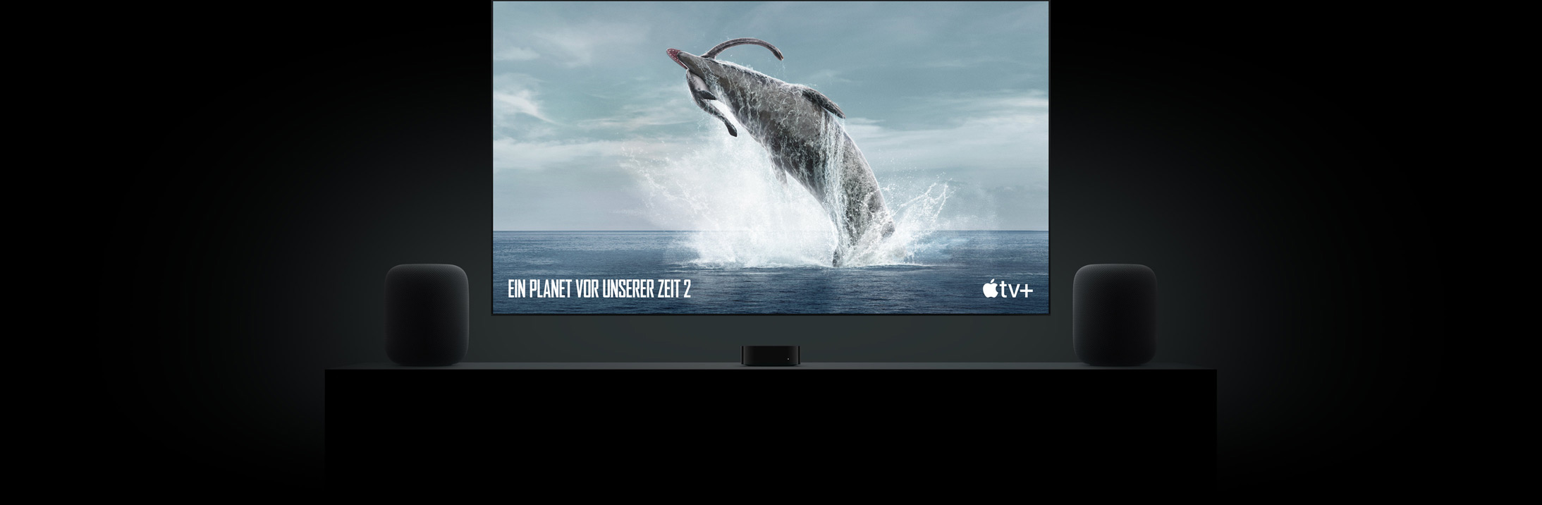 Gro?er Flatscreen Fernseher mit einem lebensechten Bild eines Dinosauriers aus ?Ein Planet vor unserer Zeit“. Der Fernseher h?ngt über einem Apple TV mit HomePod Lautsprechern links und rechts, die auf einer Wohnzimmerkonsole stehen