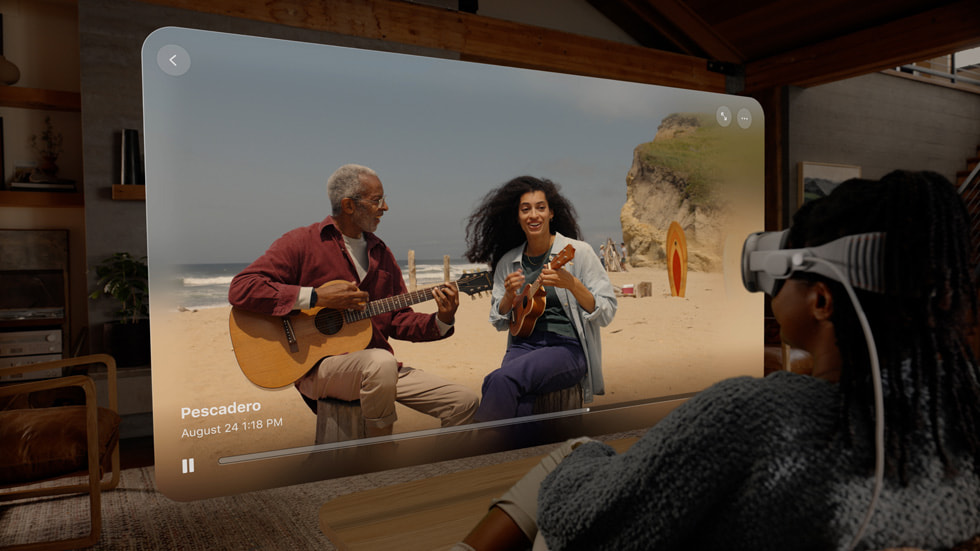 Apple Vision Pro takan biri, oturma odas?ndaki ekranda sahilde müzik yapan iki ki?inin uzamsal videosunu izliyor.