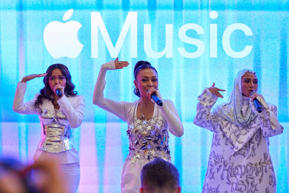 Hình ?nh c?n c?nh màn trình di?n c?a De Fam, v?i logo Apple Music trên màn hình phía sau ban nh?c. 