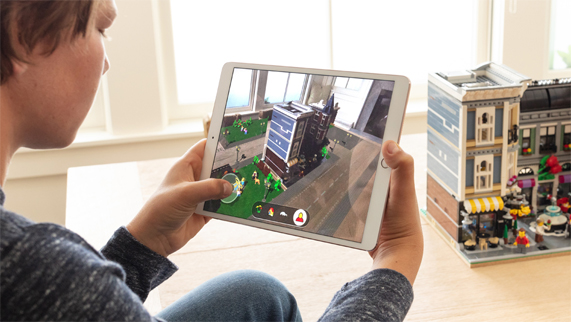 Ekran?nda LEGO AR City olan iPad’ini elinde tutarak bir odada LEGO binas?n?n ?nünde oturan ?ocuk.