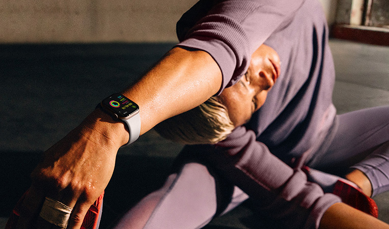 Yerde oturmu?, kolunu baca??na do?ru esneten bir kad?n?n bile?indeki Apple Watch Series 10’un ekran?nda Antrenman, So?uma, Aktivite ve Aktivite Halkalar? g?rülüyor