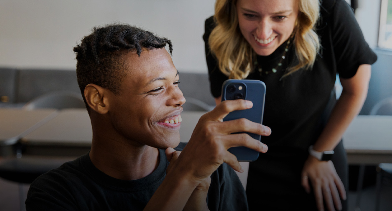 Photo d’un jeune homme avec une basse vision qui sourit et montre l’écran de son iPhone à une femme souriante, debout derrière son épaule gauche