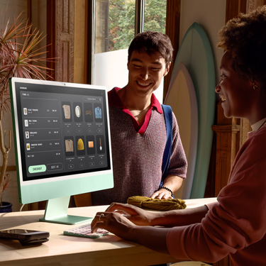 Un employé assis derrière le comptoir d’un magasin commercialisant des planches de surf et d’autres articles utilise un iMac pour finaliser une transaction avec un client se trouvant de l’autre c?té du comptoir.