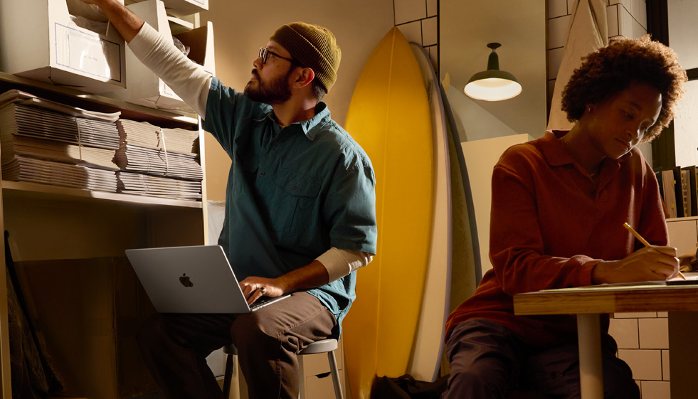 Une personne assise sur un tabouret dans un environnement de travail utilise un MacBook Air débranché posé sur ses genoux, en tendant l’autre main pour atteindre une bo?te située sur une étagère en hauteur