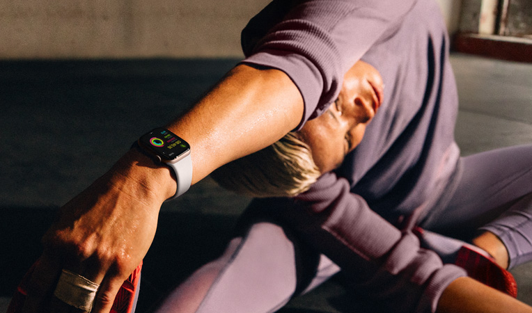 M?t ng??i ph? n? ?ang ng?i trên sàn du?i tay ch?m vào chan trong khi ?eo Apple Watch Series 10, màn hình hi?n th? Bài T?p, H? Nhi?t, Ho?t ??ng, Vòng Ho?t ??ng